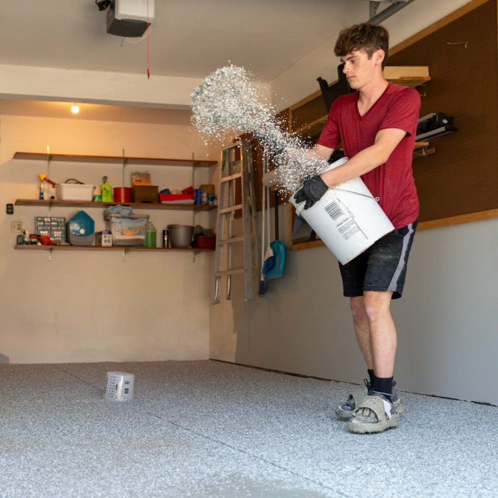 A person in a garage is spreading a substance on the floor, surrounded by tools and shelves with various items.