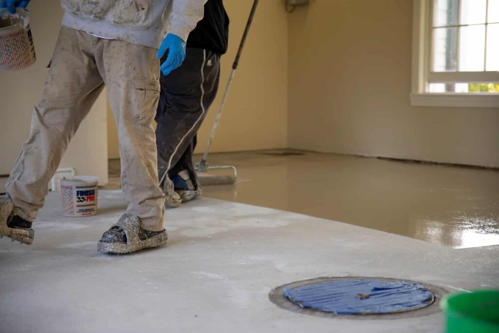 Two persons are painting a floor in a room, wearing protective gear and handling paint rollers and paint buckets.