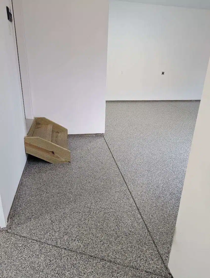 Empty, clean room with white walls and grey speckled flooring. A small wooden step is placed at the corner near a closed door.