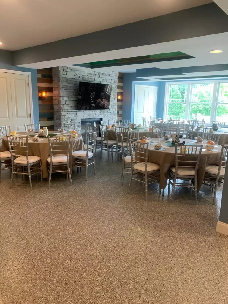 A decorated banquet room with round tables, chairs, a stone fireplace, a mounted TV, and large windows overlooking trees.