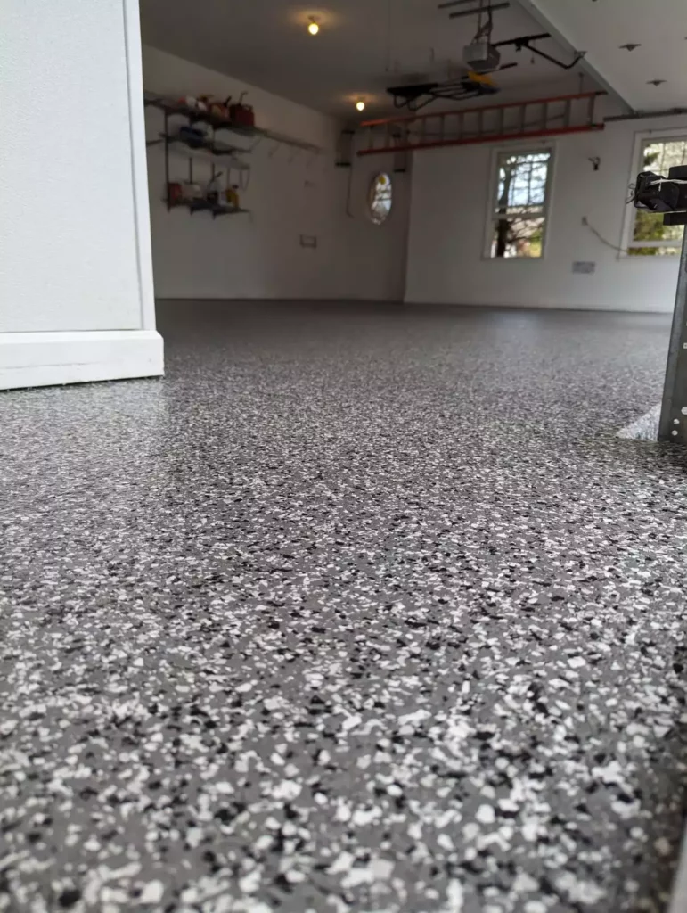 A clean garage with a speckled epoxy floor, white walls, shelving, and tools. A window and storage space are visible in the background.