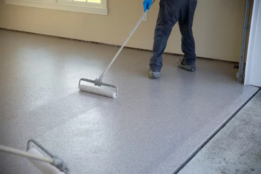 A person is applying a coating on a floor with a paint roller. The room is light-colored with a window and open doorway.